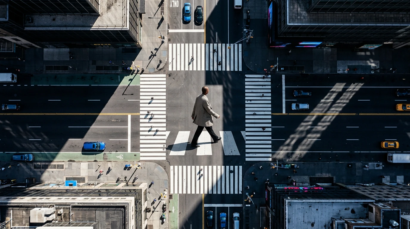 An aerial drone view looking down onto a city street, where a person walks across a crosswalk, symbolizing the two-way flow of fashion trends from runway to street style amidst geometric urban patterns.
